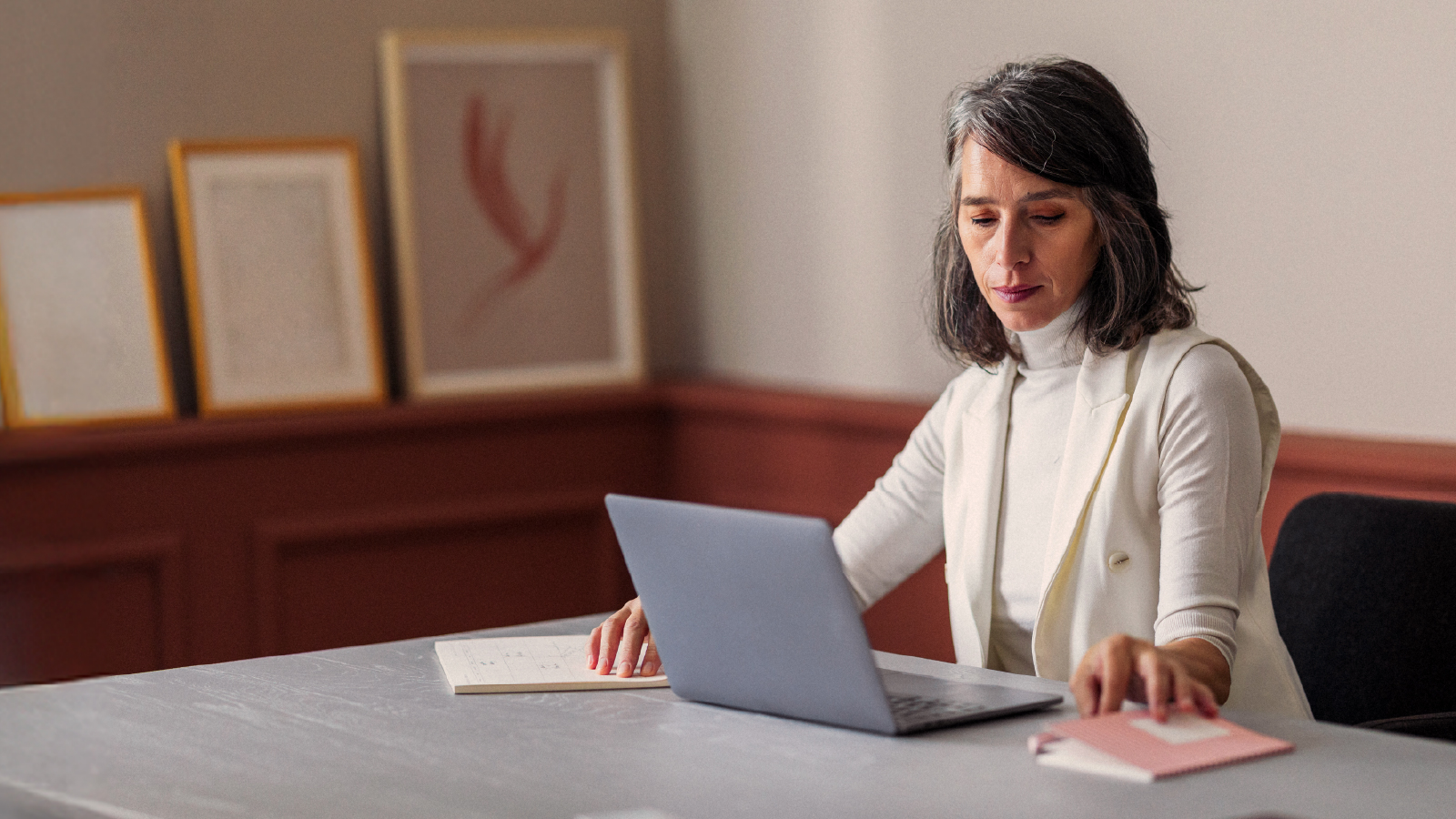 A woman sits at a desk working on a laptop, with notebooks and framed artwork in the softly lit background.