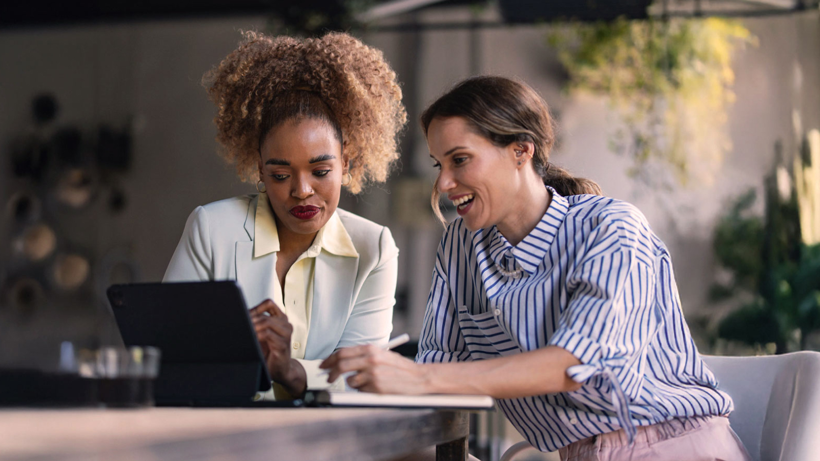 Two businesswomen sitting at a table, smiling and reviewing financial data on a tablet. The setting is a modern workspace with natural lighting and indoor plants.