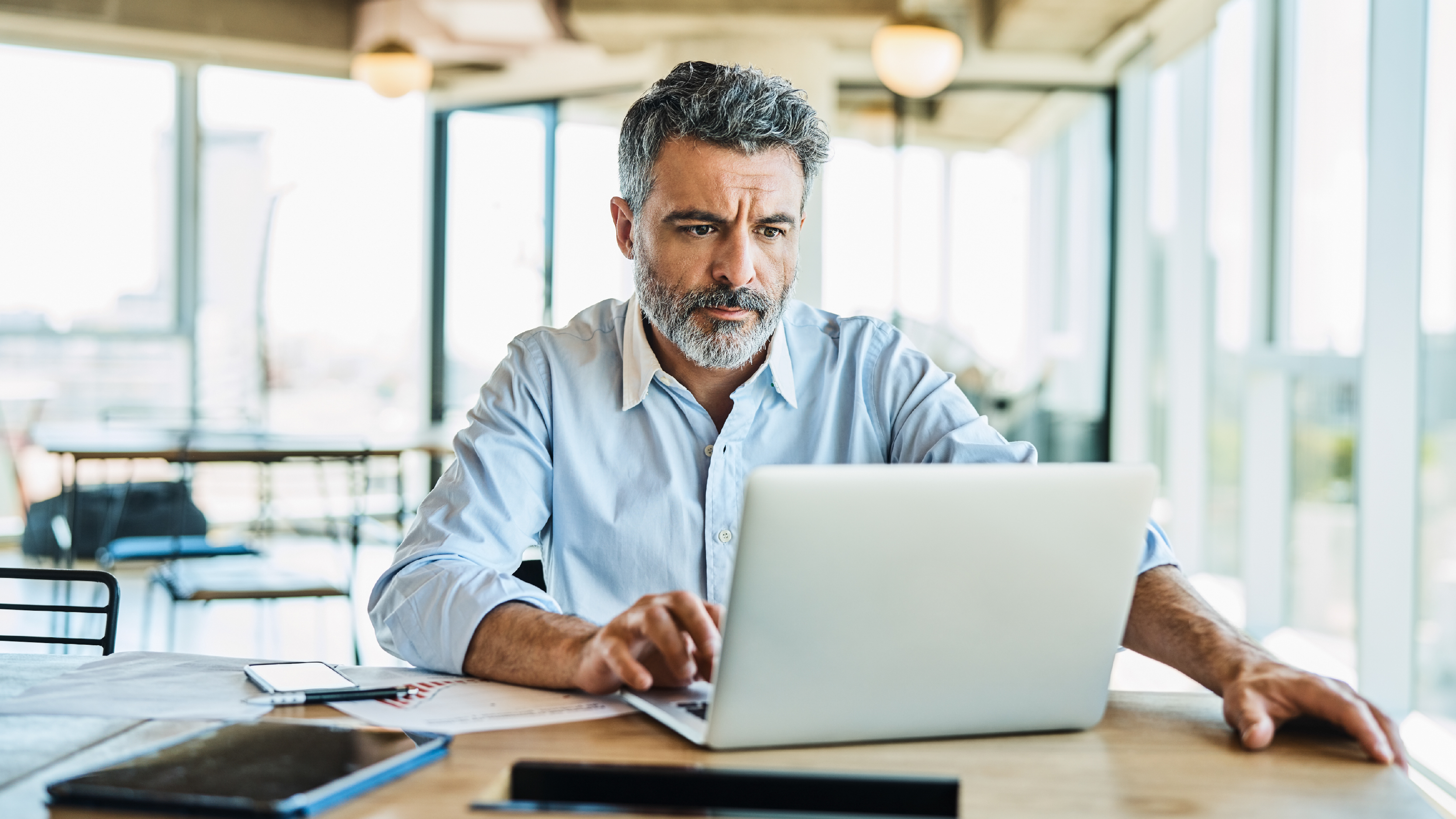 A middle-aged man with gray hair and a beard sits at a desk in a bright, open office space, looking concentrated as he types on a silver laptop. Papers, a smartphone, and a tablet lie on the desk, with large windows and modern ceiling lights visible behind him.