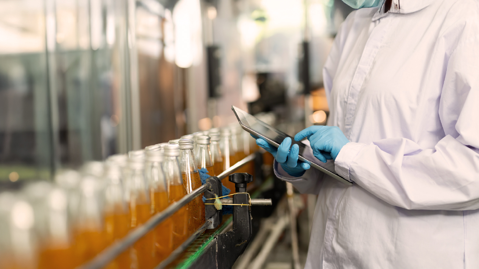 Person in a white lab coat and blue gloves using a tablet next to a production line with glass bottles in a factory setting.
