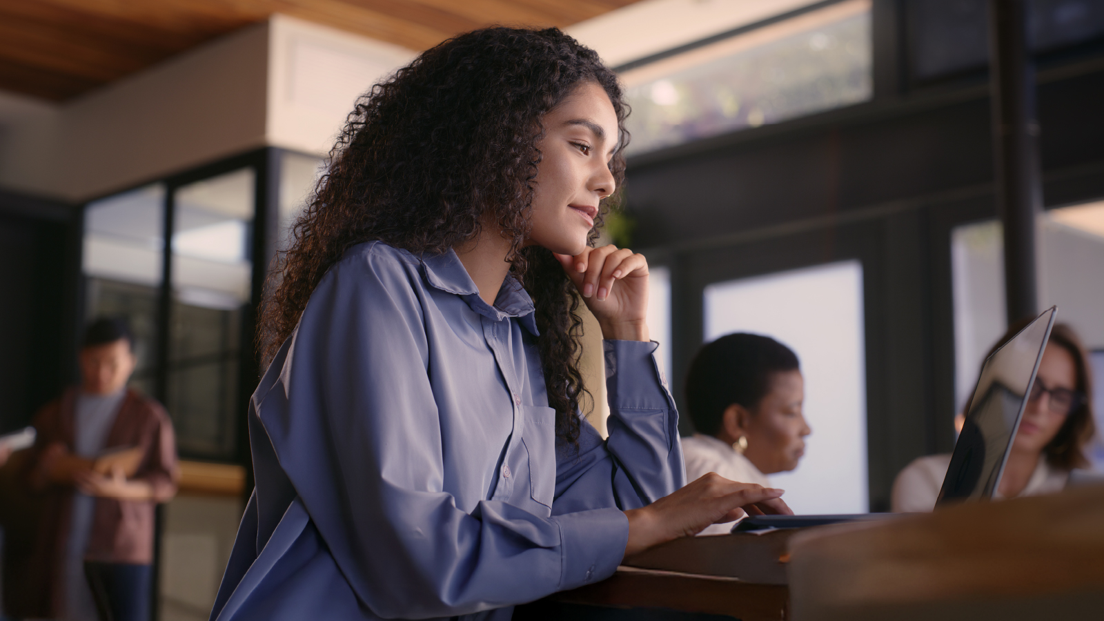 A woman in a blue shirt working at a laptop, resting her chin on her hand, focused on the screen.