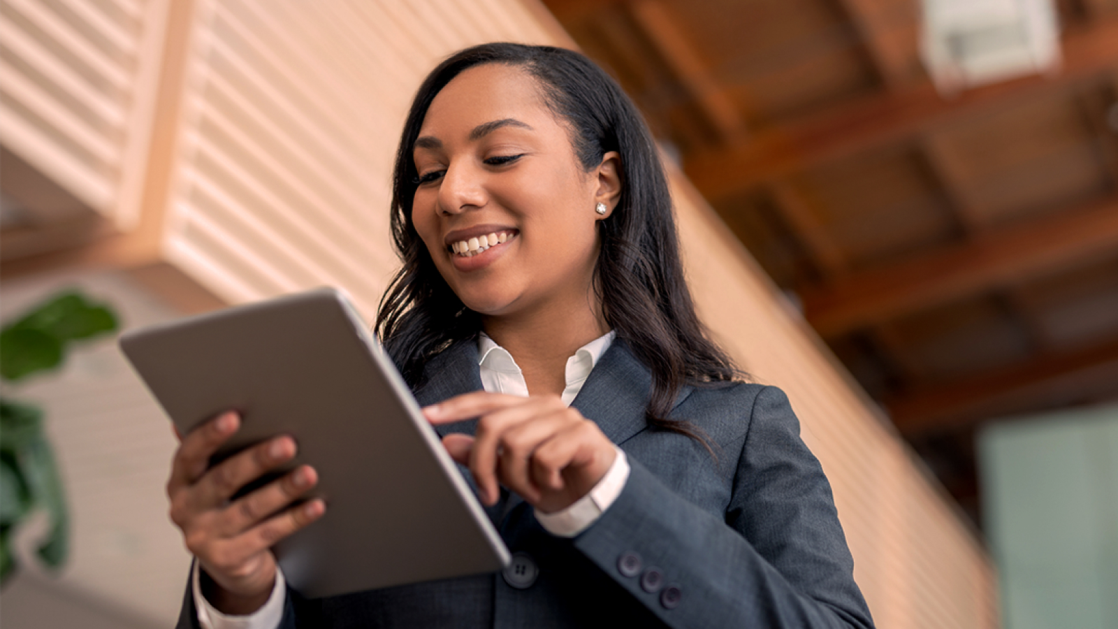 A professional woman in a business suit smiling while interacting with a tablet in a modern office environment with wooden paneling.