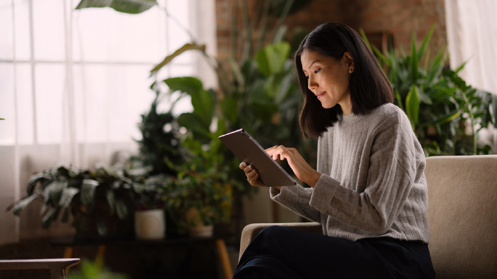 A woman wearing a gray sweater sits indoors surrounded by green plants, smiling and using a tablet.
