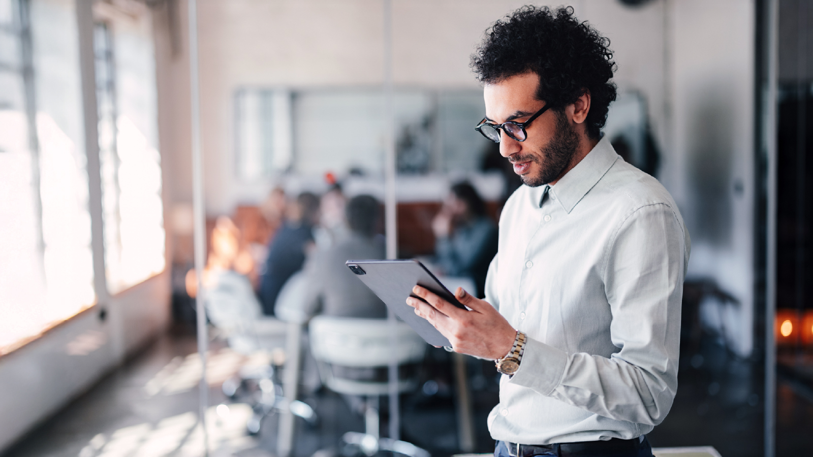 Professional standing in a bright office environment, looking at a tablet with colleagues blurred in the background.