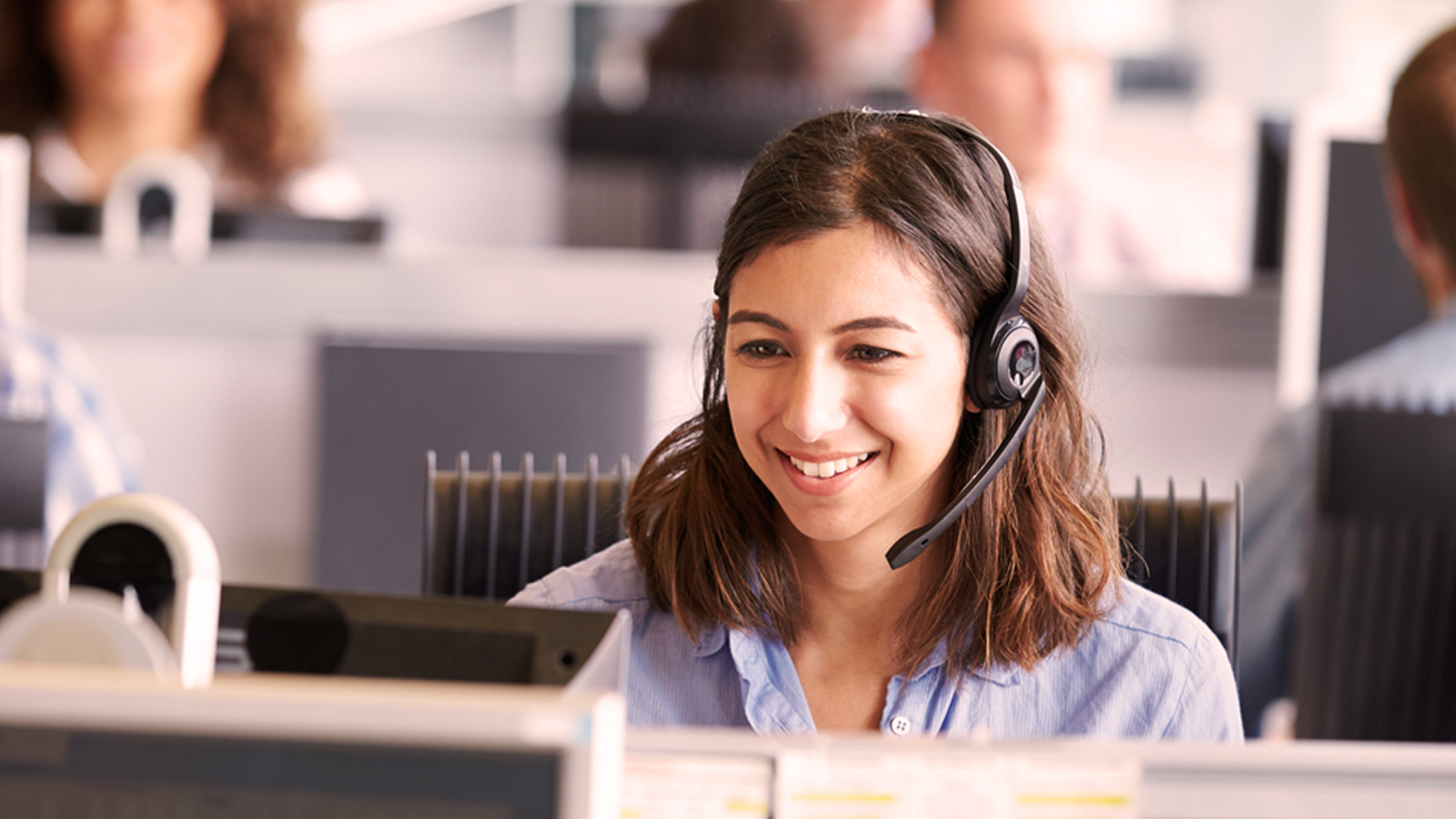 A woman smiling while wearing a headset, seated in a modern contact center environment.