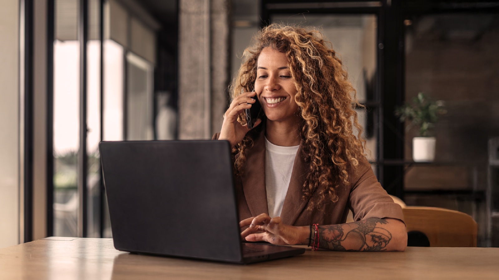 A woman with curly hair smiling while talking on the phone and working on a laptop at a wooden desk in an office setting.