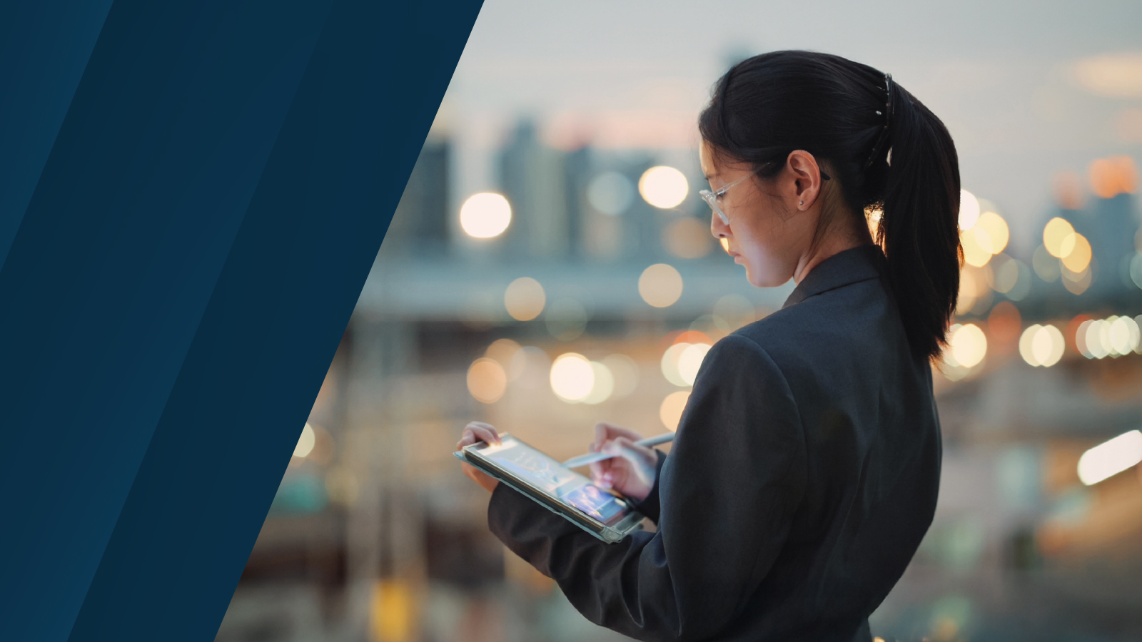 Woman in professional attire using a tablet outdoors at dusk with blurred city lights in the background.