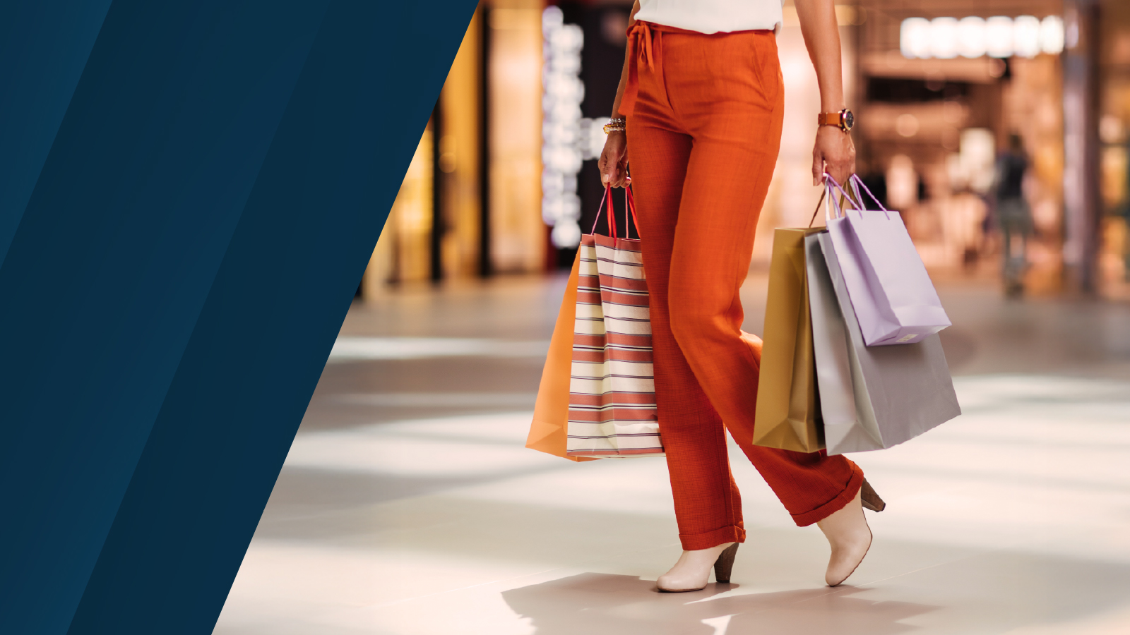 A shopper walking through a mall carrying multiple shopping bags, with a blurred retail interior in the background.
