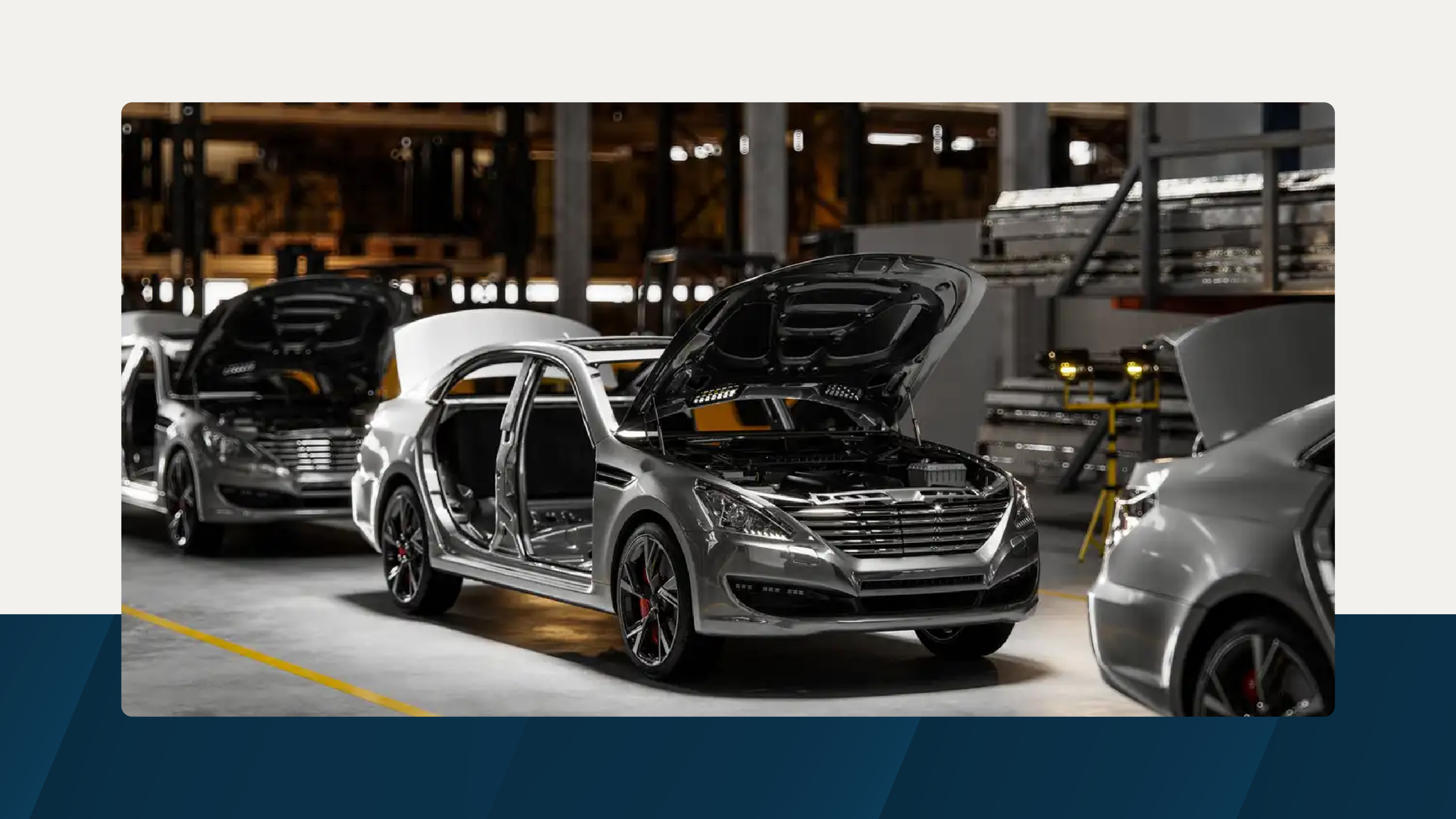 A row of partially assembled silver cars with open hoods on an automotive production line, set in a well-lit factory environment.