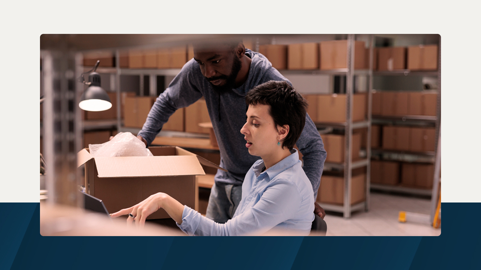 Two professionals working in a warehouse, analyzing inventory inside an open cardboard box, with shelves of packed boxes in the background.