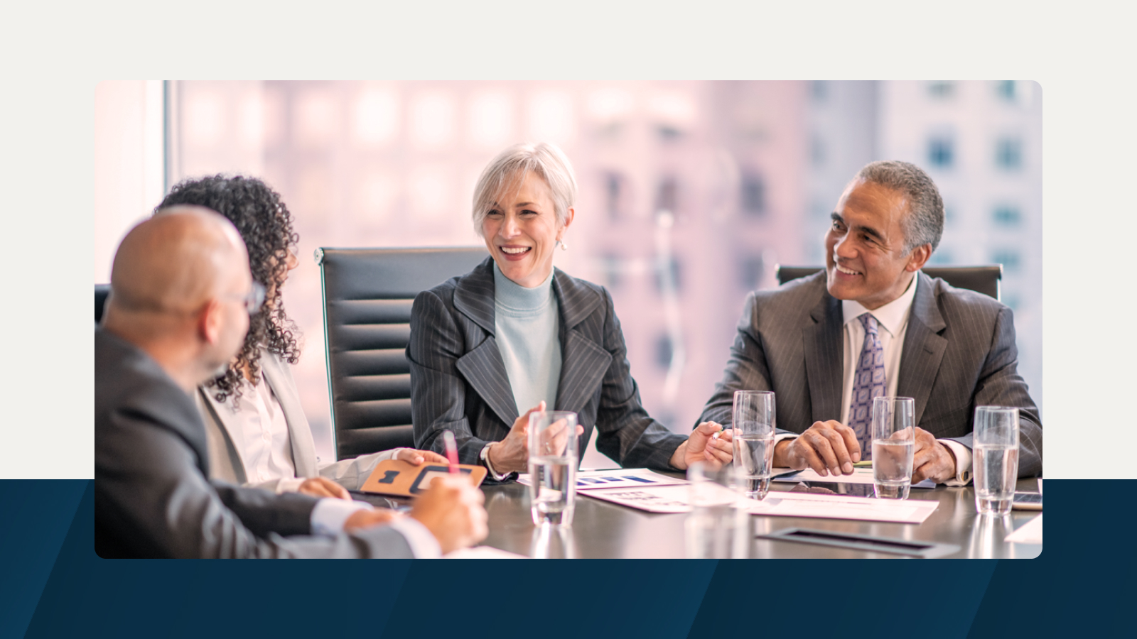 A group of professionals in formal attire seated at a conference table, engaged in a discussion and smiling, with documents and glasses of water in front of them.