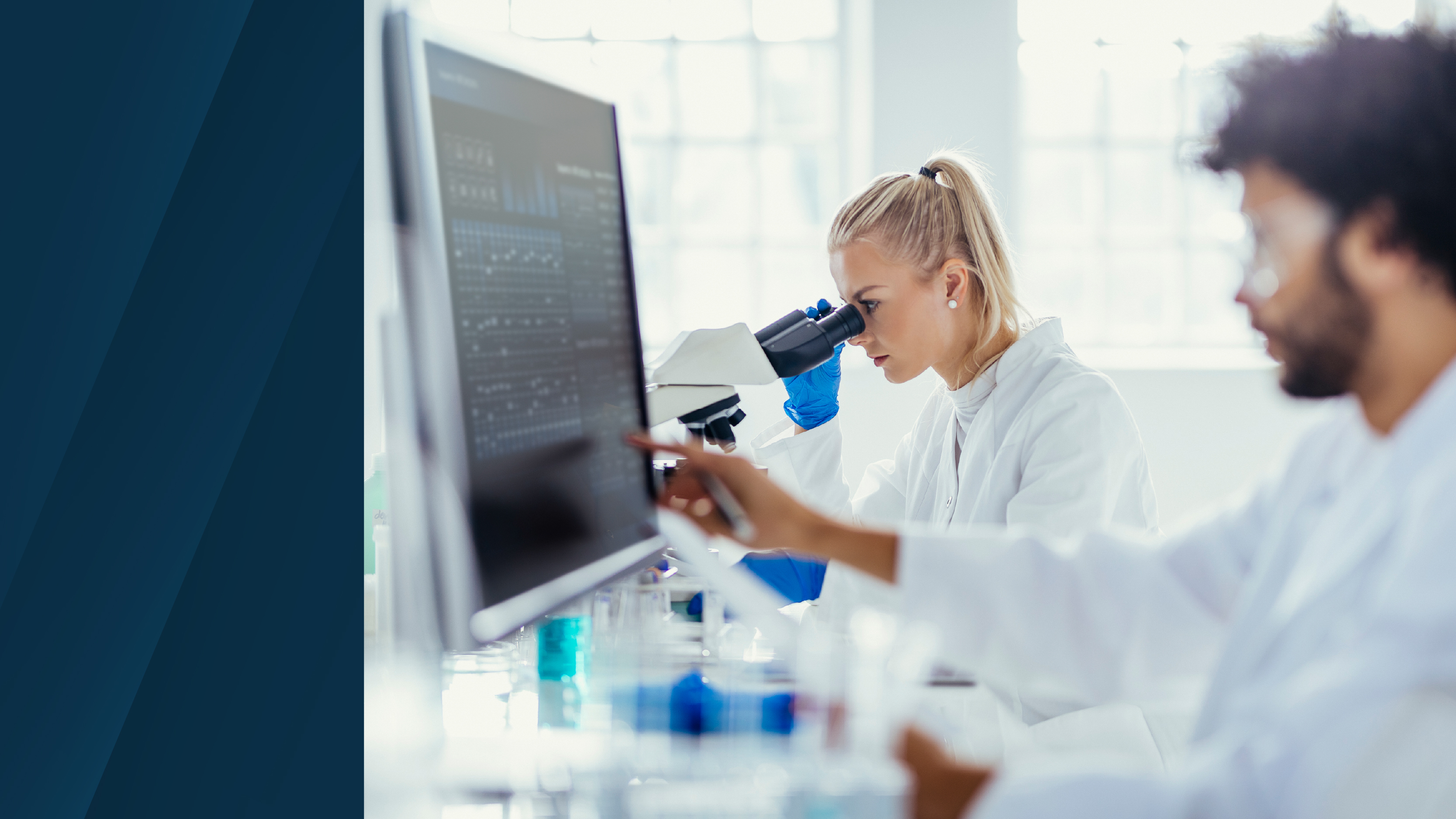 A laboratory scene showing two life sciences researchers in white lab coats reviewing data on a large monitor, with one researcher using a microscope and laboratory glassware visible in the foreground.