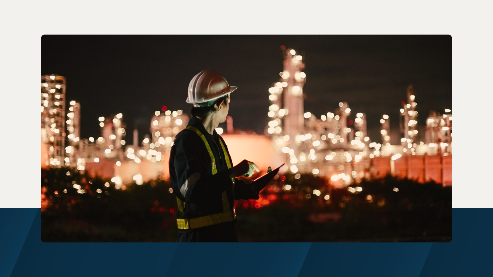 A worker in protective gear stands in front of a brightly lit refinery at night, holding a tablet and looking at the industrial infrastructure.
