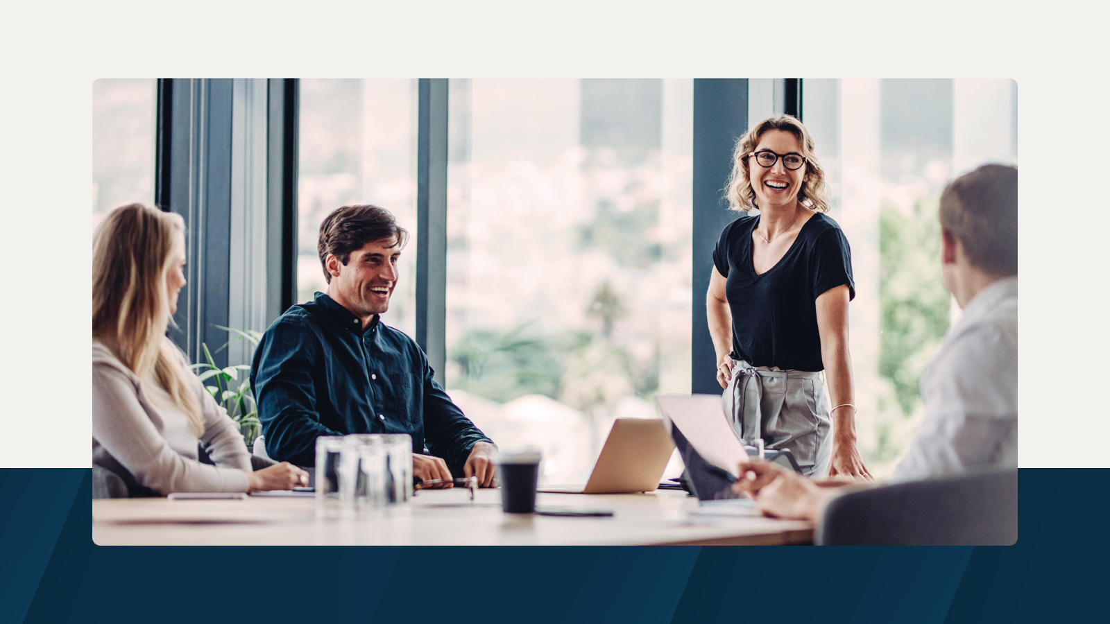 A group of colleagues in a bright meeting room, with one woman standing and smiling while others sit around a table with laptops and documents.