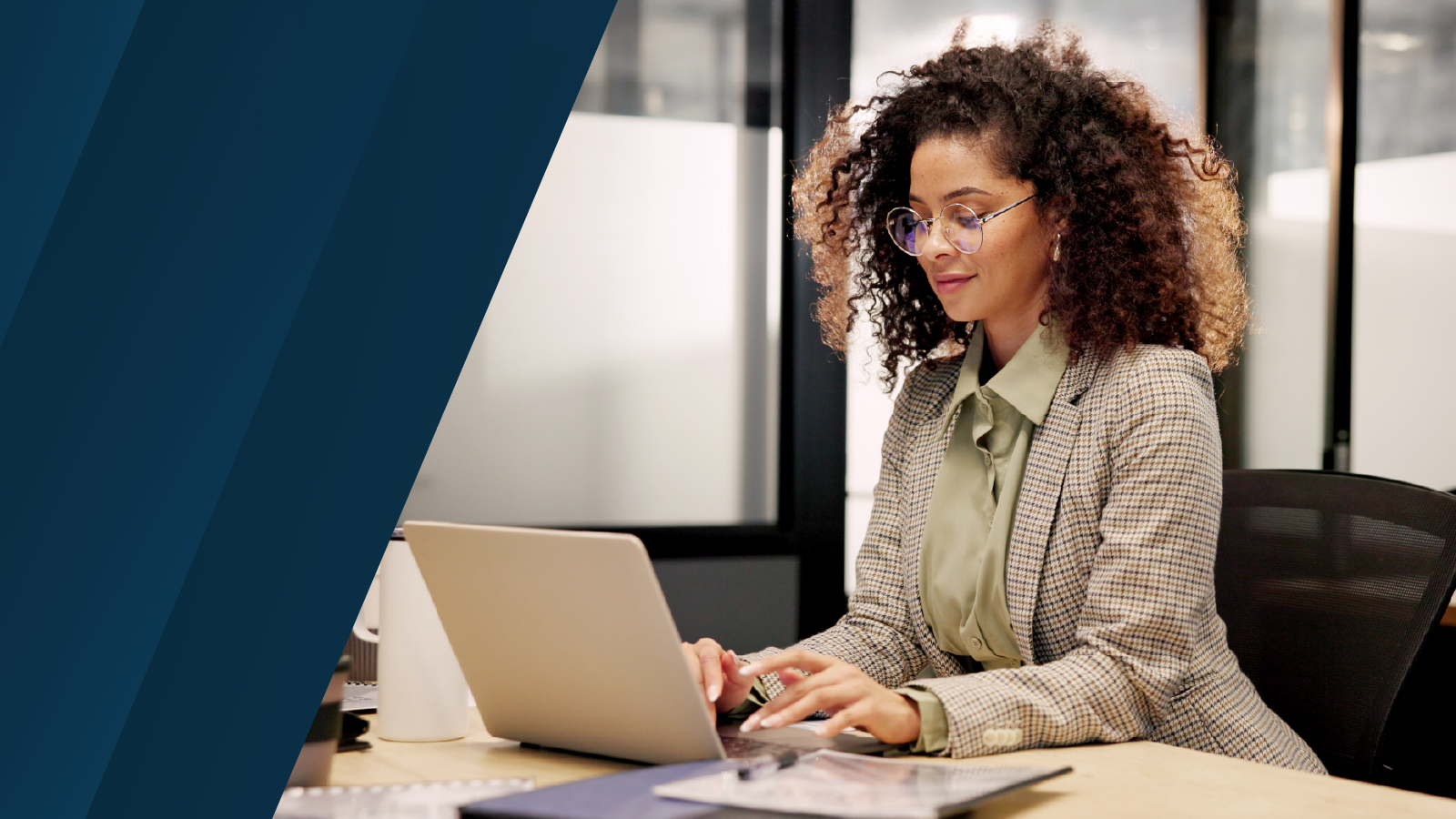 A person in a checkered blazer and green blouse sits at a desk typing on a laptop, with documents nearby in a bright, modern office environment.