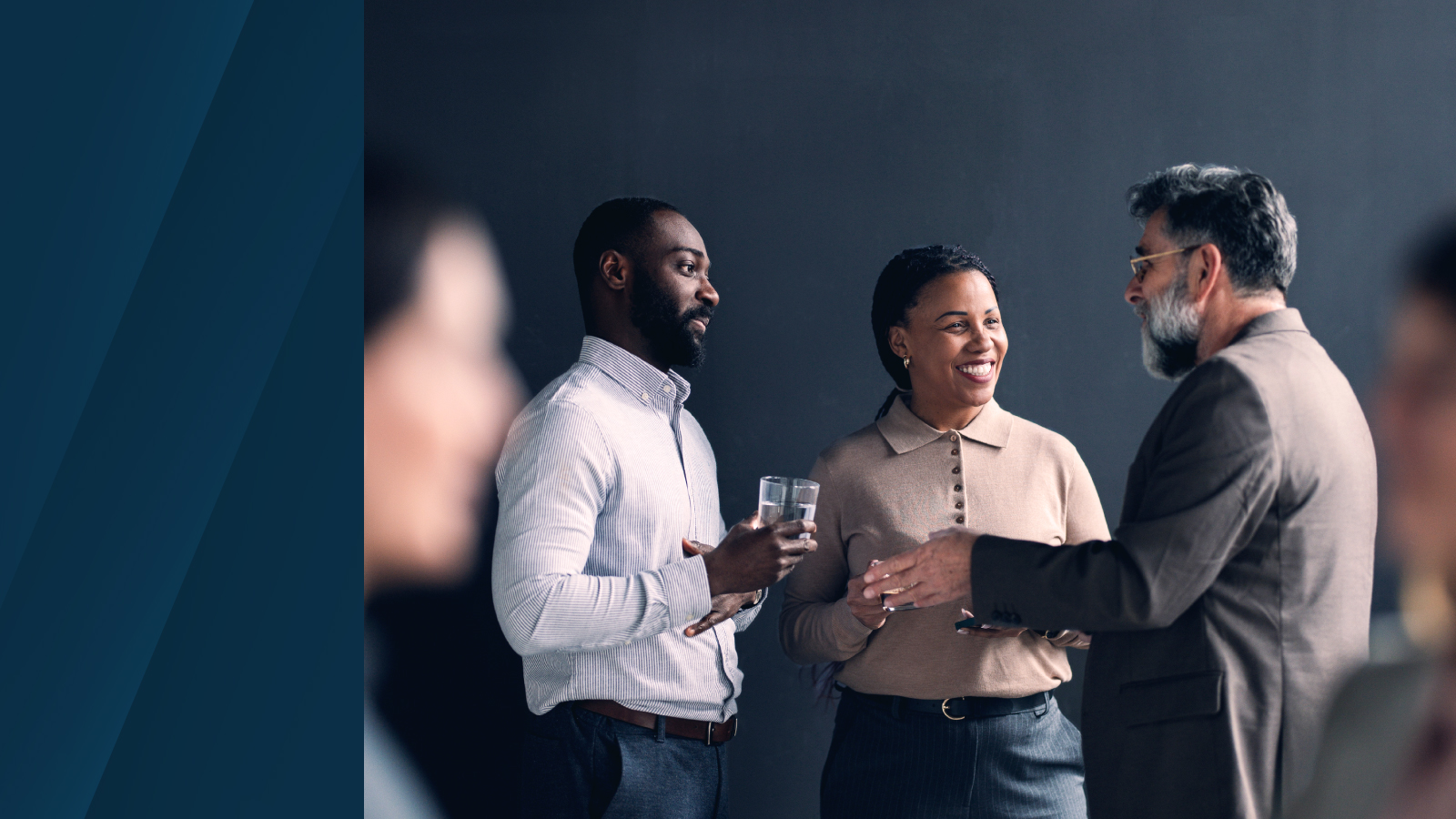 Three professionals standing and conversing in an office setting, with one person holding a glass of water, against a dark background.