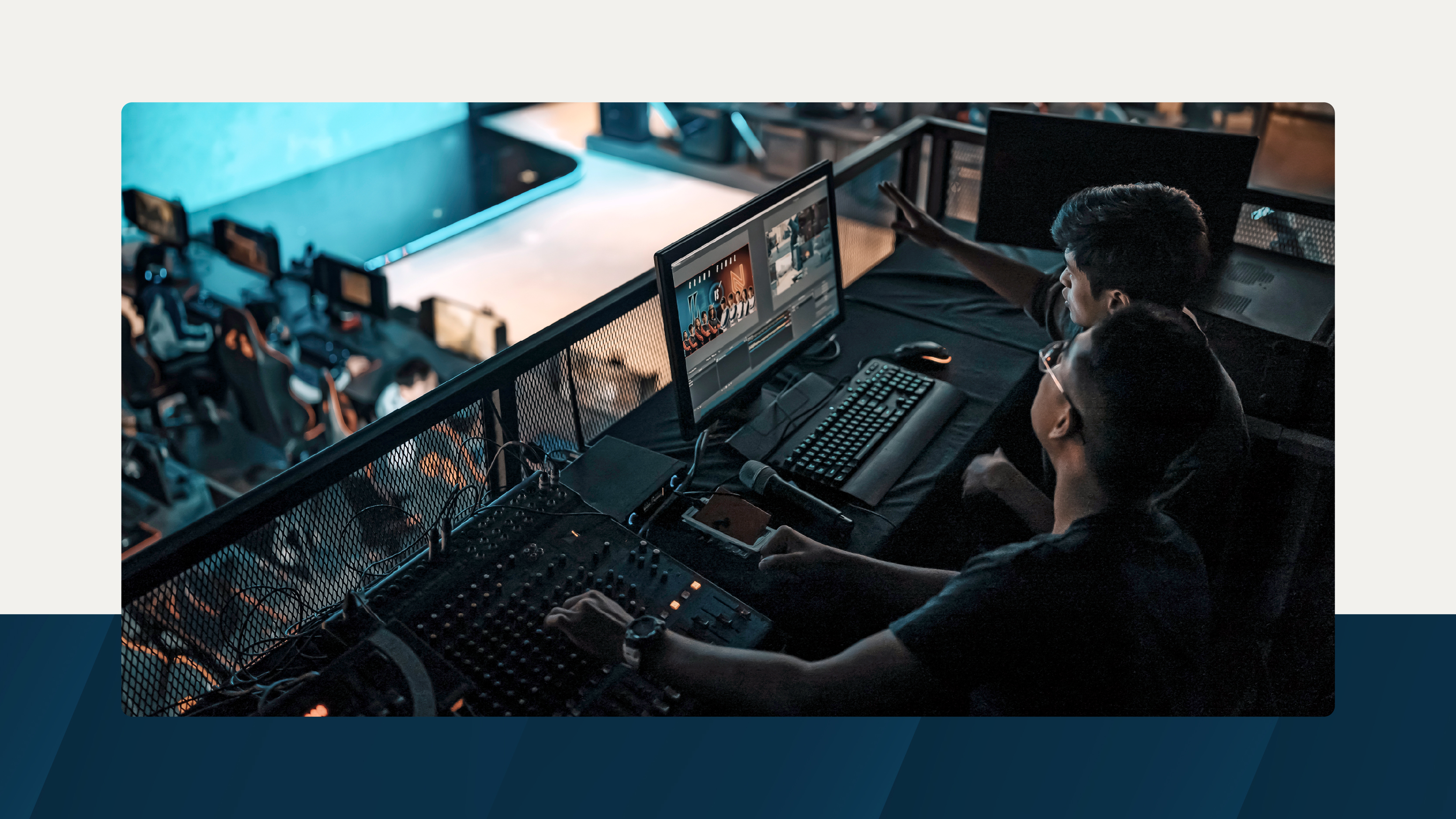 Two media professionals operate video editing and sound equipment in a production booth overlooking a room filled with computer stations and monitors, suggesting a live event or esports setting.