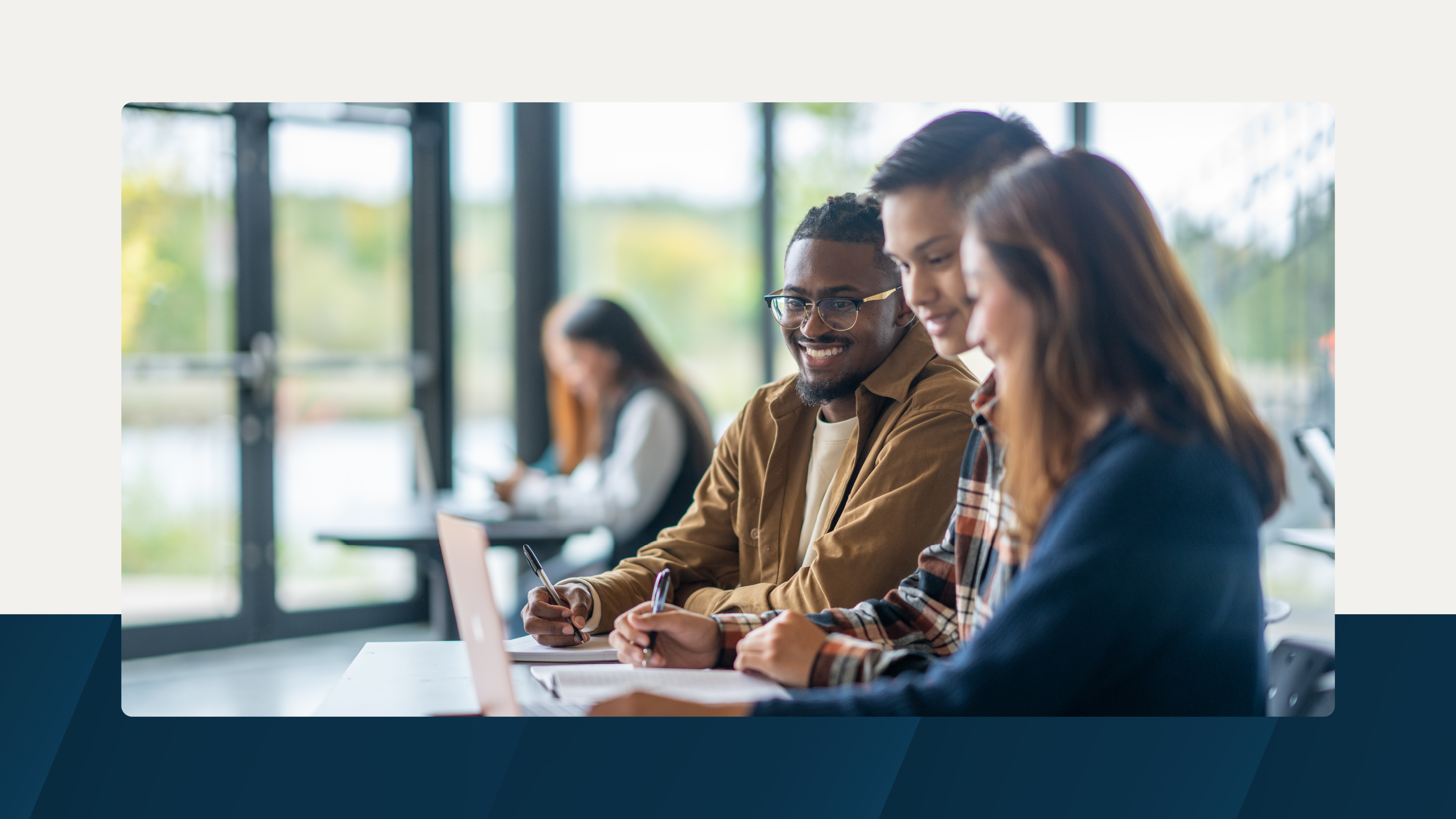 Three students collaborating with laptops and notebooks in a sunlit classroom, with large windows in the background and other students studying nearby.