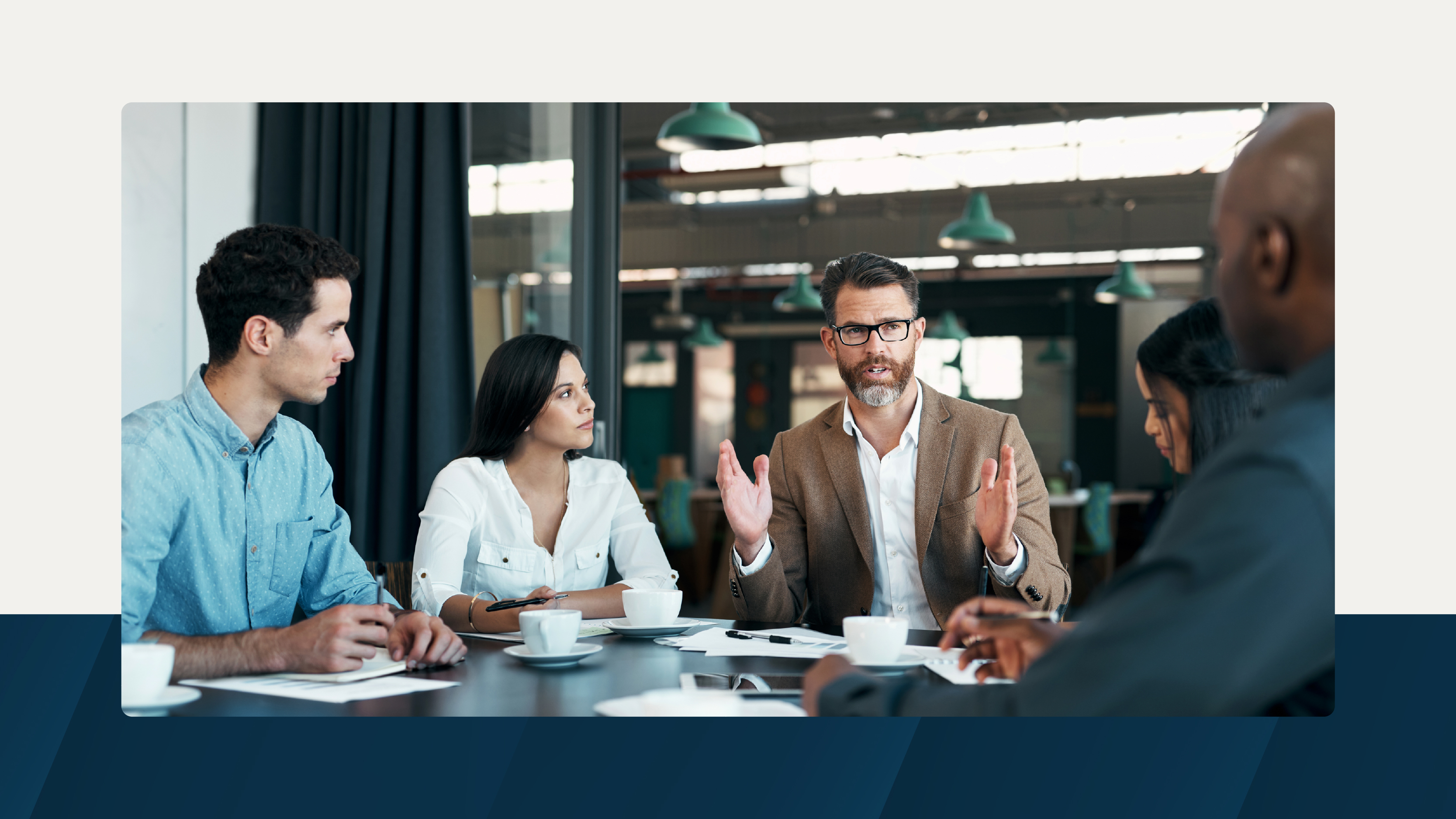 A business meeting in a modern conference room with five professionals seated around a table, actively discussing and listening.
