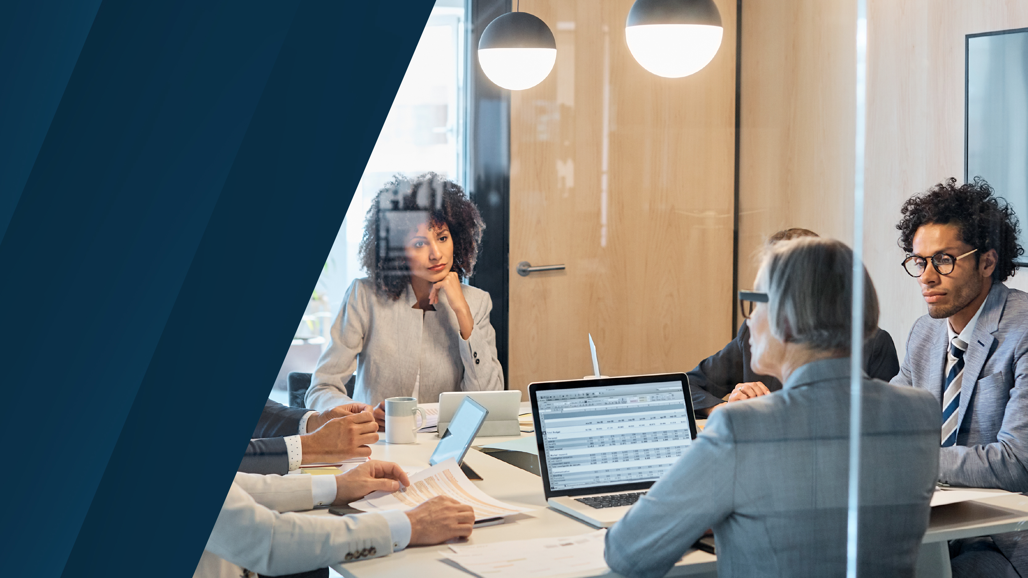 A business meeting in a modern office, viewed through a glass partition. A diverse group of professionals in formal attire discuss financial data displayed on a laptop. A blue design element is on the left side.