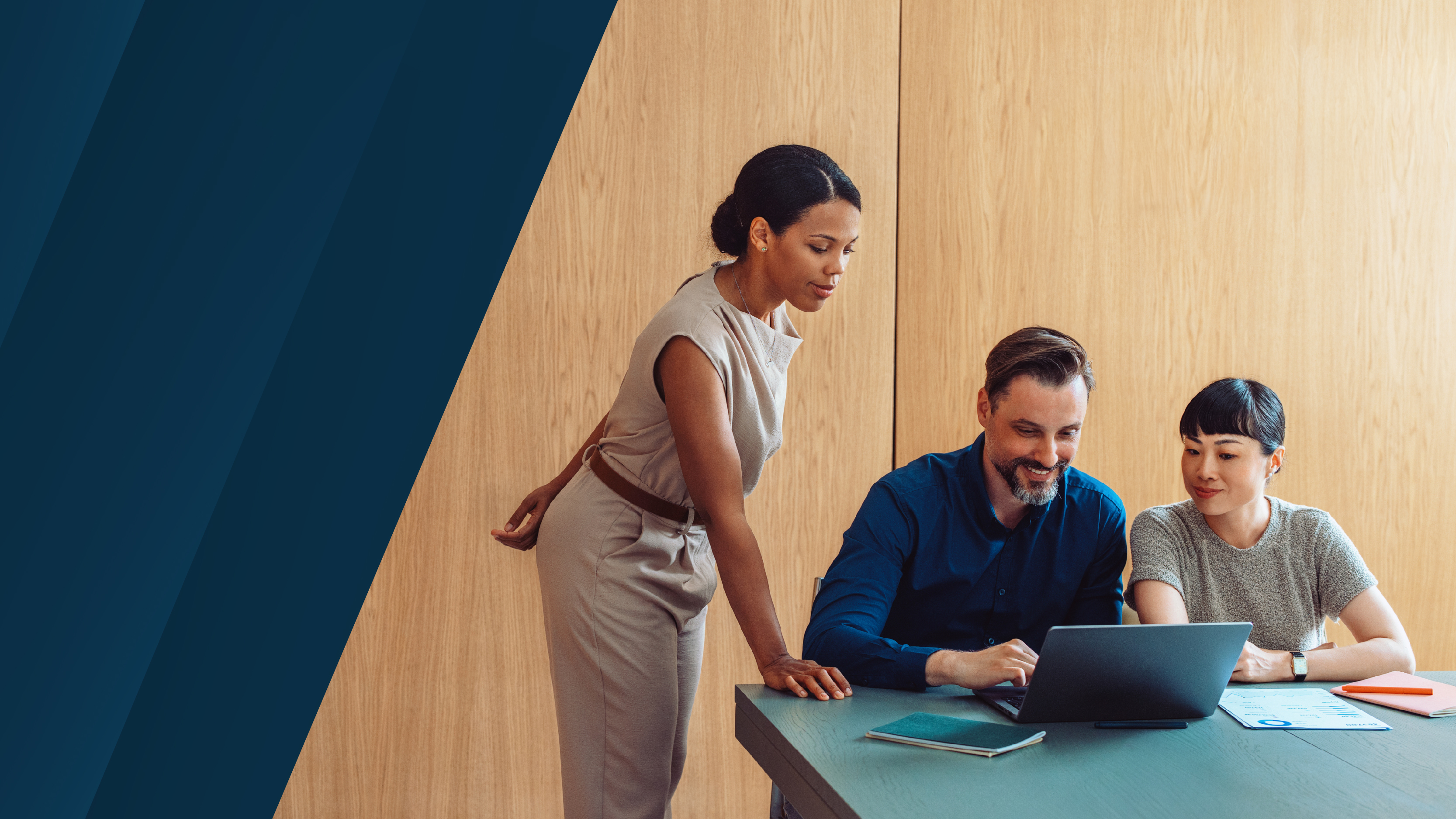 A woman stands beside two colleagues seated at a table, all focused on a laptop screen as they review documents together in a bright meeting room with wood paneling.