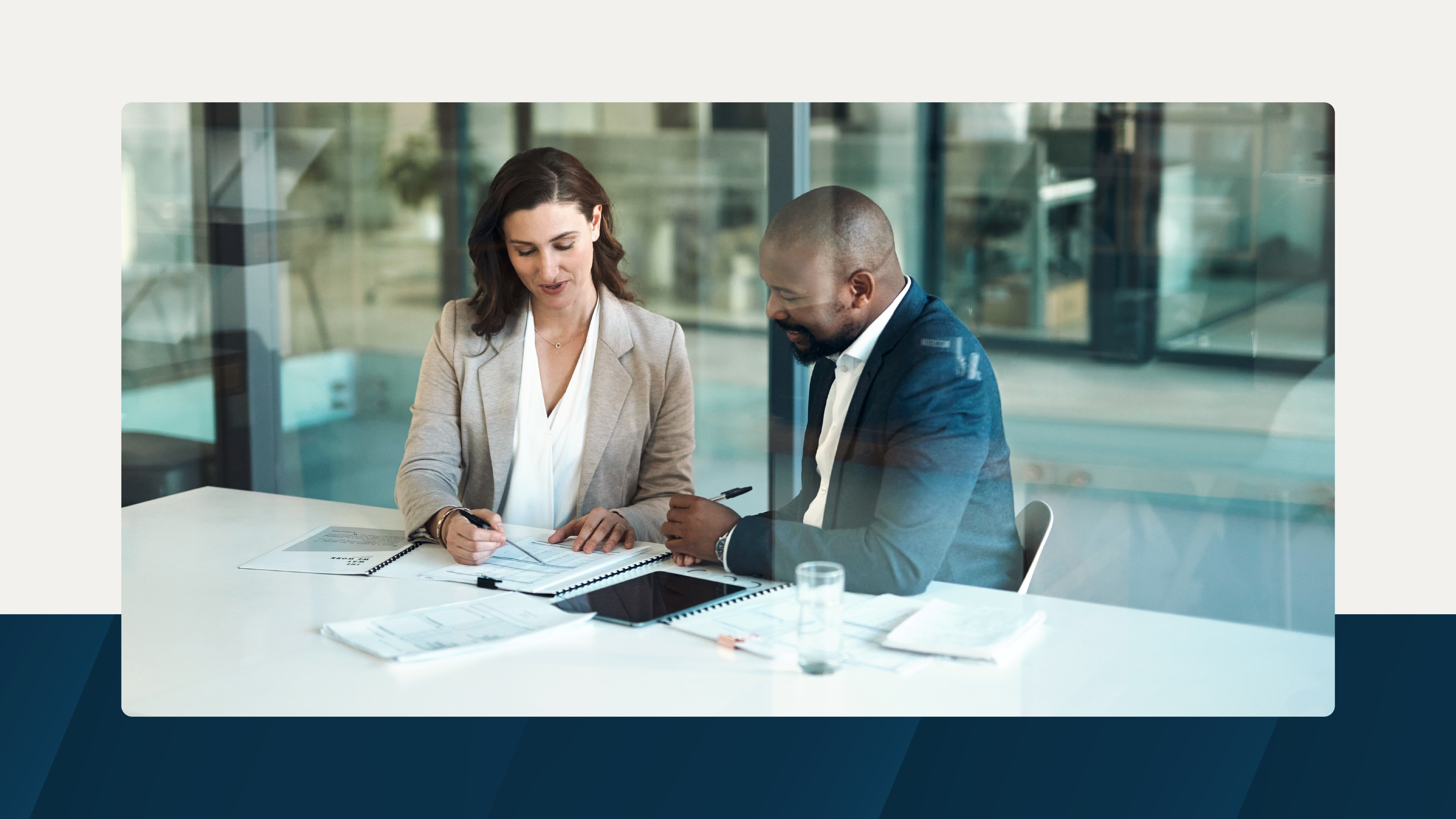 Two business professionals in formal attire collaborate at a conference table with documents and a tablet in a glass-walled office.