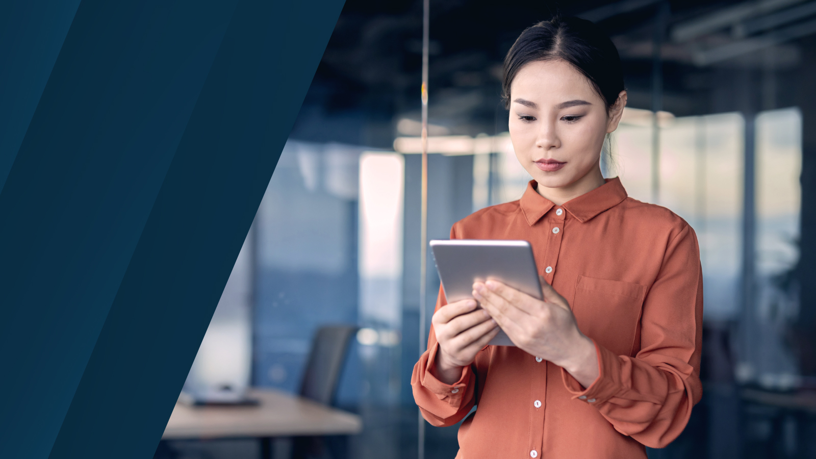 A woman in an orange blouse is focused on using a tablet in a modern office setting with glass walls and a blurred background. A dark blue ink design is present on the left side of the image.