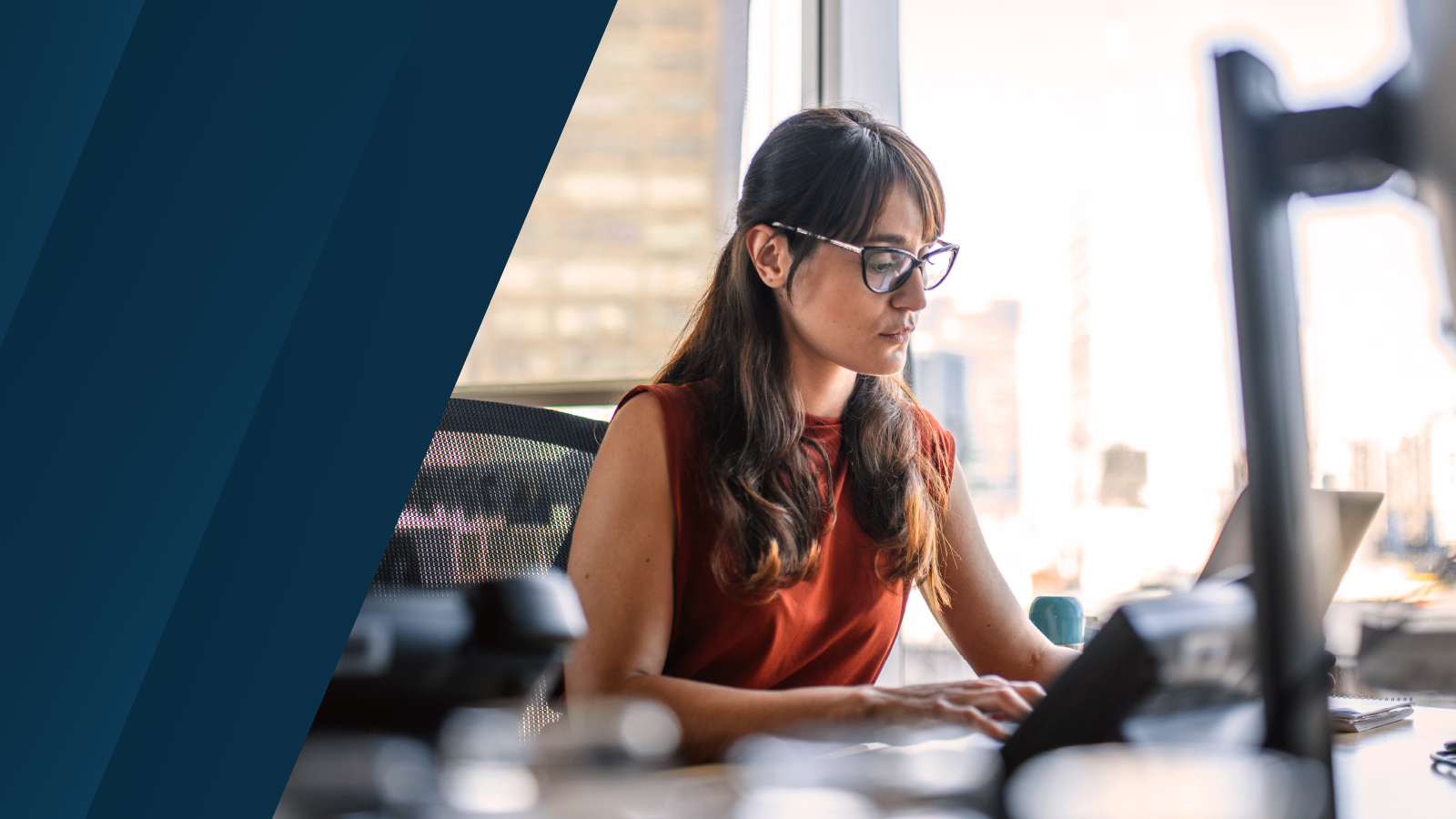 A woman in glasses sits at a desk working on a laptop, focused on the screen, with office items blurred in the foreground.