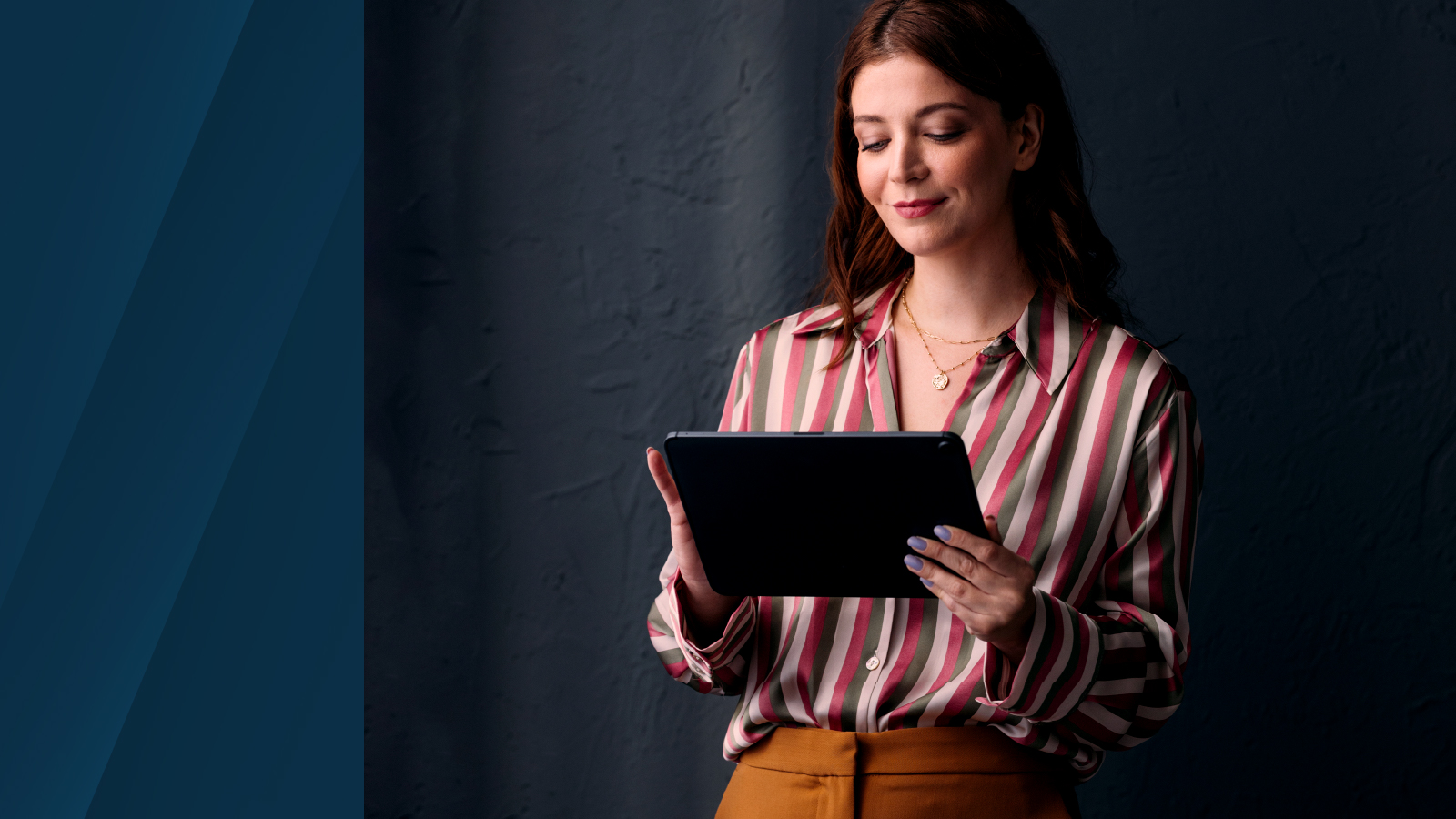 A woman in a striped blouse and mustard pants holding a tablet, standing against a dark textured wall with a blue background panel on the left.