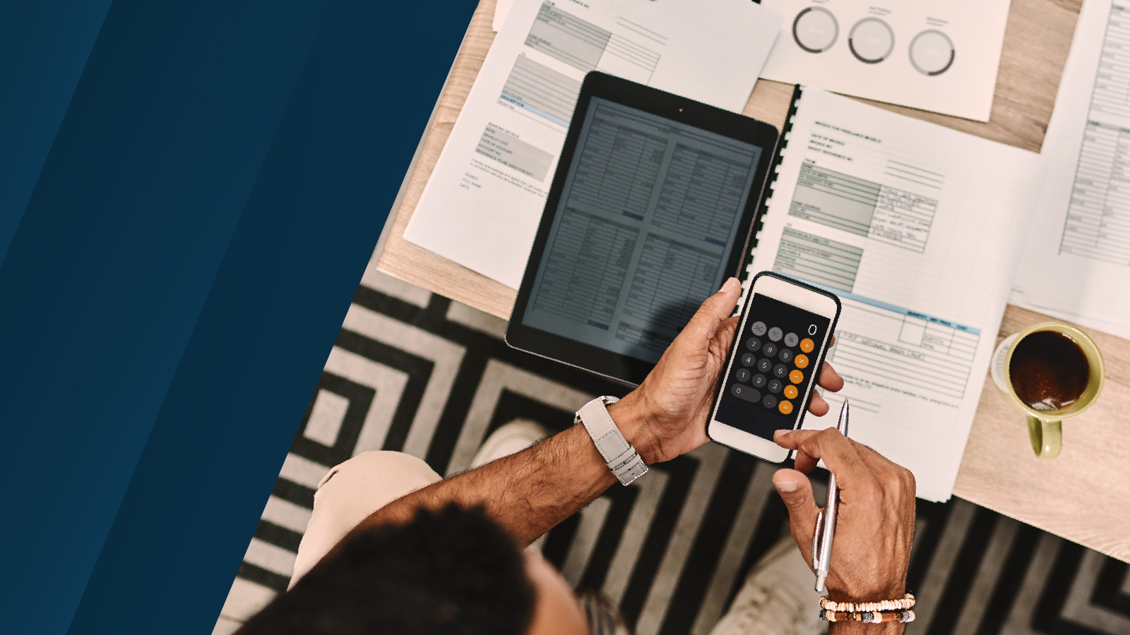 Overhead view of a person using a smartphone calculator app while reviewing financial documents and a digital tablet, placed on a desk with a striped carpet below.