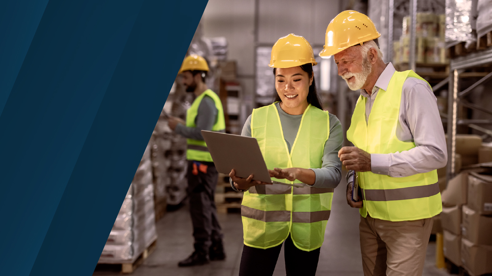 Two workers in yellow safety vests and helmets standing in a warehouse aisle, smiling while reviewing information on a laptop.