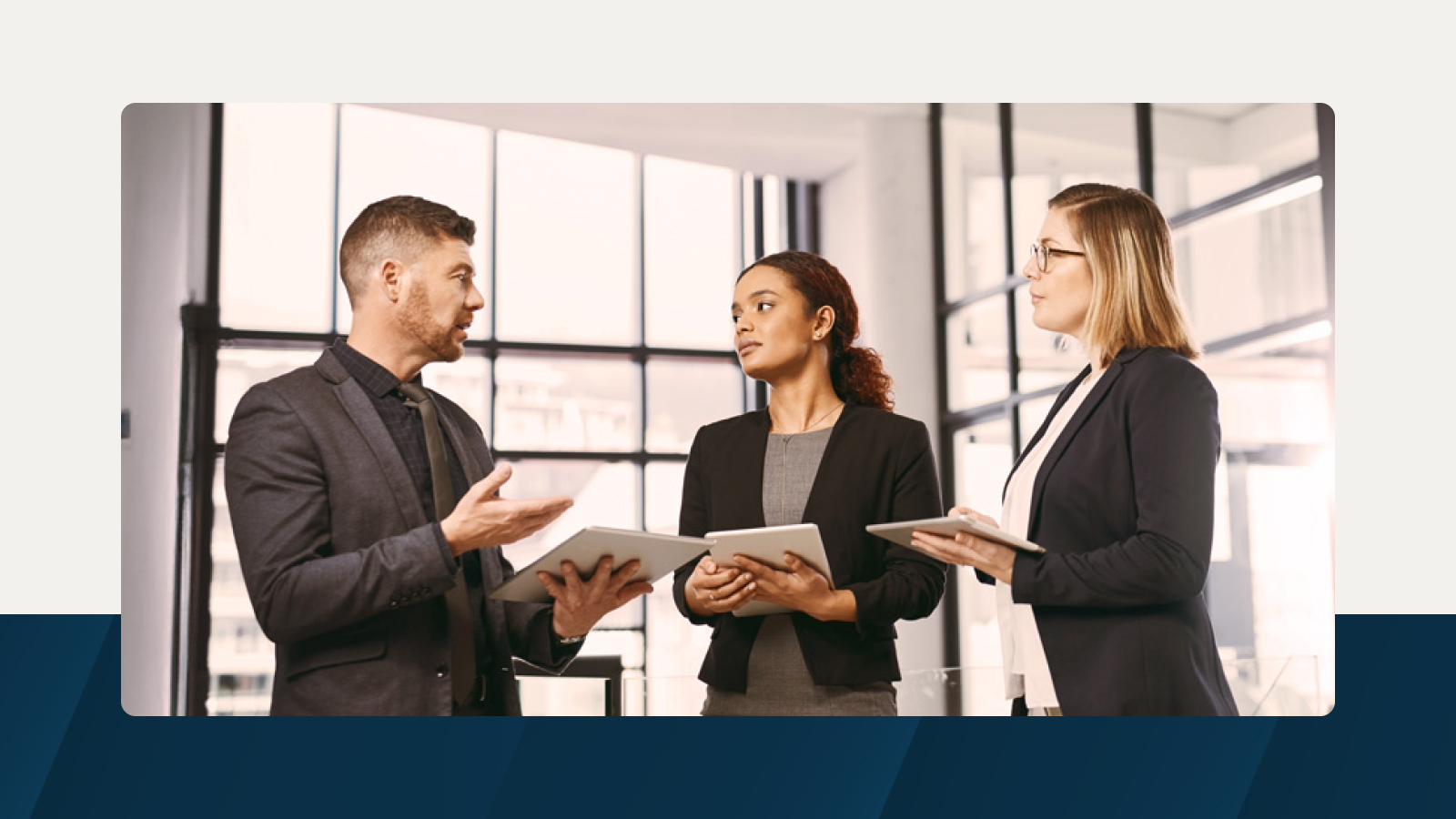 Three business professionals in formal attire discuss operating expense planning while holding tablets in a modern office with large windows.
