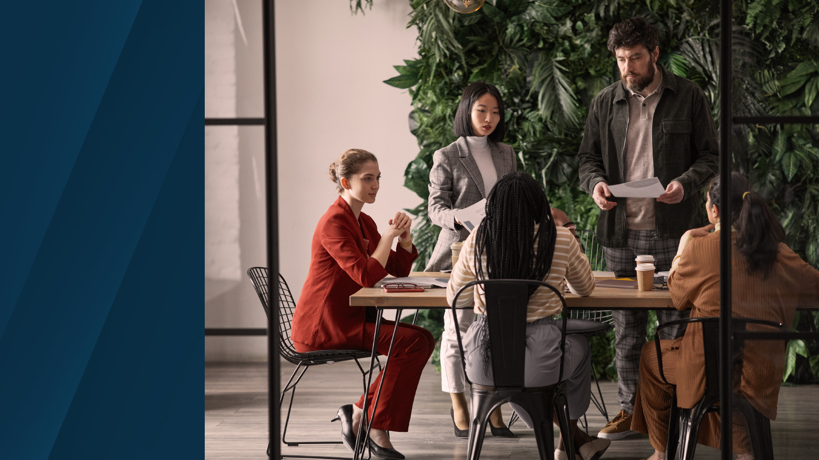 Group of professionals in business attire collaborating around a meeting table, reviewing documents and discussing strategies. Green foliage wall in the background adds a natural setting.